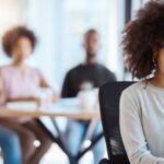 Medewerker voelt zich buitengesloten op de werkvloer, kijkt verdrietig terwijl collega's praten in de achtergrond.